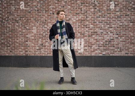 Un jeune homme se tient en toute confiance dans la ville pendant l'automne, portant un manteau noir élégant et une écharpe colorée. La toile de fond du mur de briques ajoute une touche de U. Banque D'Images