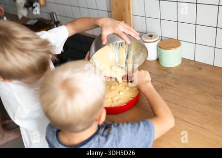Deux enfants préparent avec impatience une pâte à gâteau dans une cuisine confortable, versant le mélange dans un bol rouge vif. Leur travail d'équipe et leur plaisir soulignent le Banque D'Images