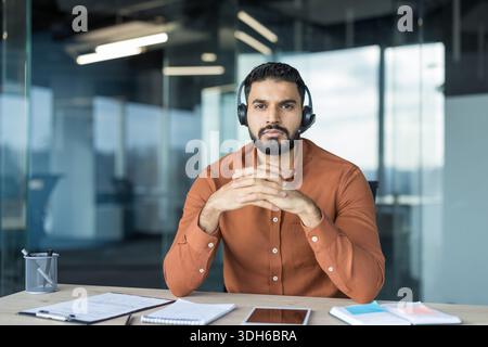 Jeune homme portant un casque professionnel avec un microphone, assis à un bureau moderne avec les mains serrées, regardant directement, prêt pour une réunion en ligne ou un appel au service client Banque D'Images