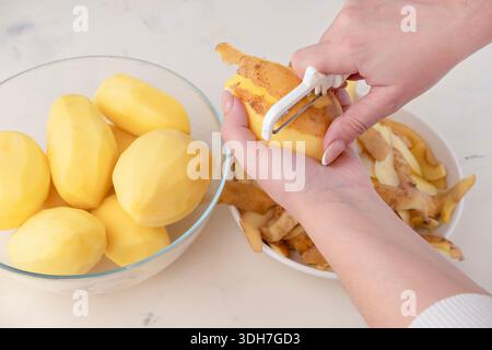 Mains d'une femme au foyer épluchant la pomme de terre crue avec un couteau d'outil spécial gros plan, vue de dessus. Banque D'Images