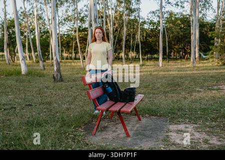 Jeune femme aux cheveux rouges debout derrière un banc rouge de parc avec un sac à dos placé sur le siège. Banque D'Images