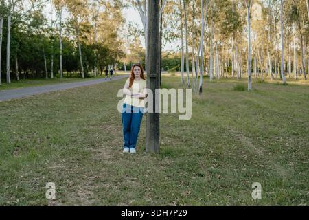 Plan large d'une jeune femme aux cheveux roux se tenant en partie cachée derrière un arbre dans un parc public. Banque D'Images