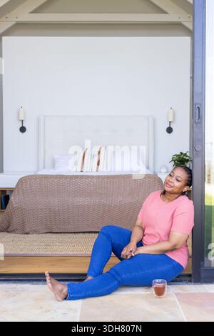 Femme afro-américaine assise sur le seuil de porte coulissante à la chambre à coucher à côté de tasse de thé, espace de copie Banque D'Images
