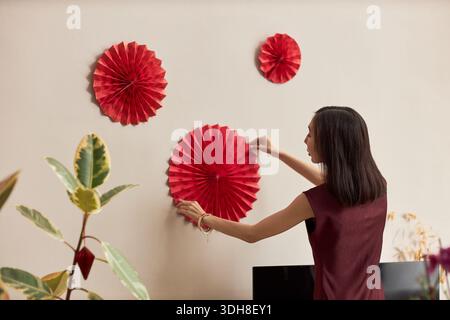 Jeune femme adulte asiatique décorant le mur avec des ventilateurs en papier rouge pour la célébration des fêtes, debout à l'intérieur et arrangeant des décorations festives pour Têt, plantes visibles au premier plan Banque D'Images