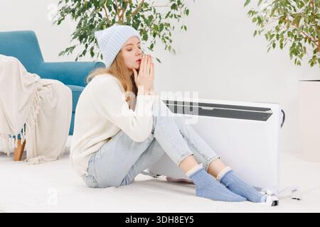 Jeune femme dans des vêtements chauds se trouve sur le sol par chauffage électrique portable dans la maison froide pendant l'hiver économisant l'énergie Banque D'Images