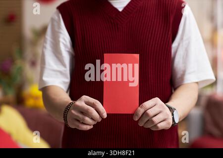 Jeune homme adulte asiatique tenant une enveloppe rouge avec les deux mains devant la poitrine, portant une chemise à manches courtes et un pull sans manches, célébrant la fête traditionnelle du Têt à l'intérieur Banque D'Images