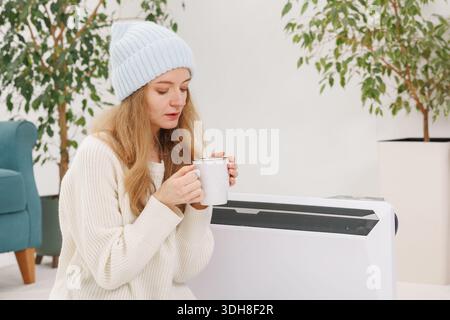 Femme habillée chaude tient une tasse de boisson chaude à côté d'un radiateur électrique portable dans un appartement froid avec espace de copie pour le contenu de coût d'énergie Banque D'Images