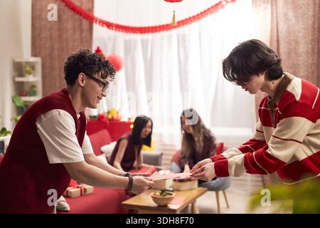 Jeune homme asiatique adulte remettant un cadeau à la jeune femme asiatique adulte, tandis que deux jeunes femmes asiatiques adultes assis en arrière-plan pendant la célébration des vacances dans le salon décoré Banque D'Images