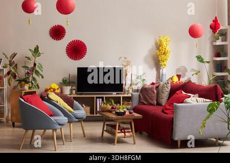 Salon décoré avec des lanternes rouges, des ornements festifs et des fleurs jaunes pour la célébration de vacances, table basse affichant la nourriture traditionnelle, télévision sur support en bois Banque D'Images