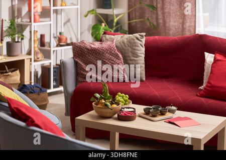 Salon décoré pour les vacances Tet avec table en bois avec plateau de fruits, service à thé, enveloppes rouges et collations, canapé avec coussins rouges et décorations festives en arrière-plan Banque D'Images