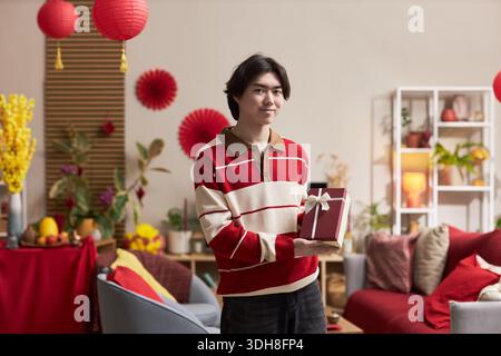 Portrait de jeune homme asiatique adulte souriant et tenant une boîte-cadeau, tout en se tenant debout dans le salon décoré avec des ornements de vacances Tet et des plantes festives visibles en arrière-plan Banque D'Images
