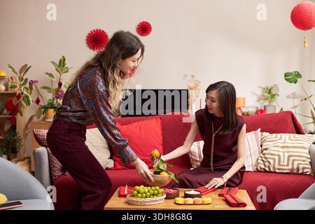 Jeune femme asiatique adulte arrangeant des fruits sur la table, tandis qu'une autre jeune femme asiatique adulte assise sur le canapé atteignant la nourriture pendant la célébration des vacances Têt dans le salon décoré avec des ornements rouges Banque D'Images