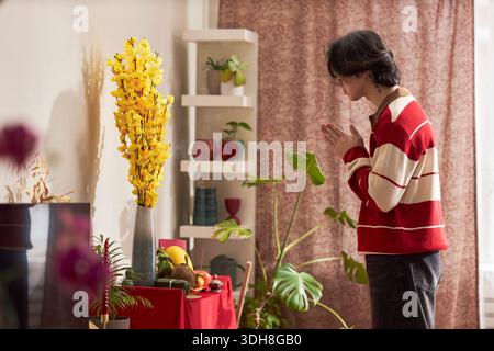 Jeune homme asiatique adulte debout avec les mains pressées ensemble dans un geste de prière devant la table de l'autel, décoré avec des fleurs jaunes et des fruits pendant la célébration des vacances du Têt à la maison Banque D'Images