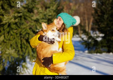 Jeune femme en veste chaude tient corgi dans la neige au parc d'hiver montrant l'amour des animaux de compagnie et le style de vie en plein air confortable Banque D'Images