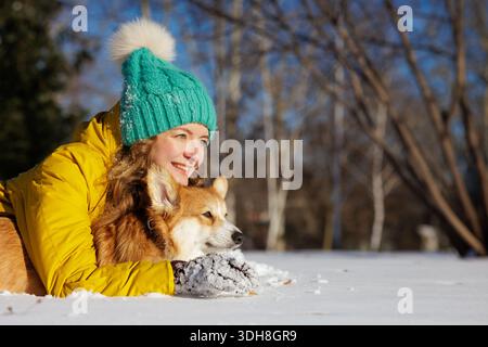 Femme souriante dans la veste chaude embrasse le chien corgi sur la neige dans le parc d'hiver pendant la promenade en plein air avec grand espace de copie Banque D'Images