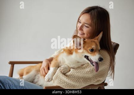 Heureuse jeune femme câlin corgi sur le fauteuil à la maison avec espace de copie montrant le style de vie et les soins détendus des propriétaires d'animaux de compagnie Banque D'Images