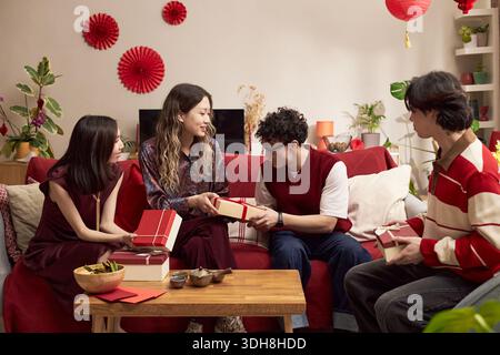 Groupe de jeunes hommes et femmes asiatiques adultes échangeant des cadeaux, assis sur un canapé dans le salon décoré avec des ornements rouges et des lanternes pendant la célébration des vacances Banque D'Images