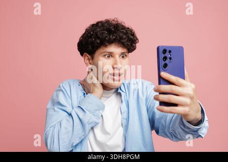Jeune homme aux yeux larges choqué par un message inattendu sur smartphone pendant l'appel vidéo isolé sur fond rose Banque D'Images