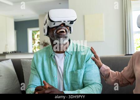 Couple senior afro-américain assis sur le canapé à la maison, homme portant un casque VR riant Banque D'Images