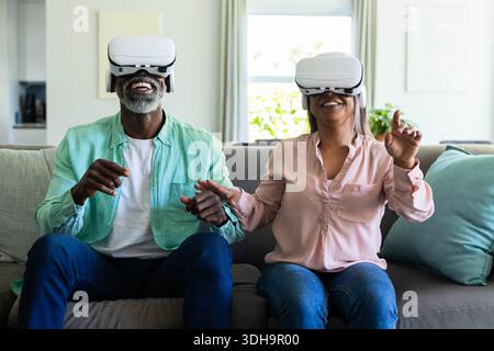 Couple diversifié portant des casques VR blancs, assis et faisant des gestes sur un canapé gris dans un salon lumineux Banque D'Images