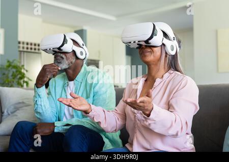Couple senior portant des casques VR blancs assis sur un canapé dans le salon interagissant virtuellement Banque D'Images