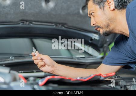 Vue latérale d'un bel homme asiatique avec barbe change de bougies d'allumage dans sa voiture. Remplacement des bougies d'allumage dans la voiture. Banque D'Images