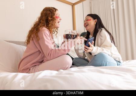 Diverses femmes amies riant, tenant du vin et un smartphone sur le lit dans des vêtements de détente, des patchs hydrogel Banque D'Images