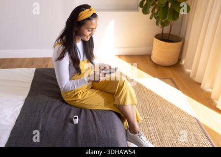 Femme asiatique assise sur le lit dans la chambre, dans une combinaison jaune tenant smartphone, appareil blanc Banque D'Images