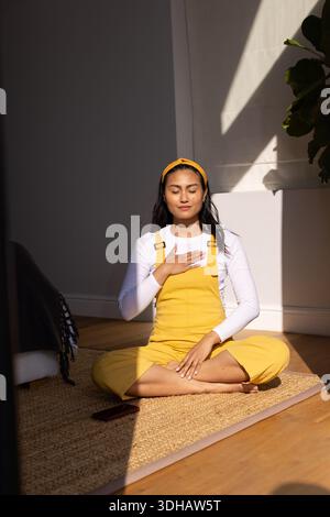 Femme asiatique adulte moyenne assise sur un tapis dans une pièce ensoleillée dans une salopette jaune, méditant près d'un smartphone Banque D'Images