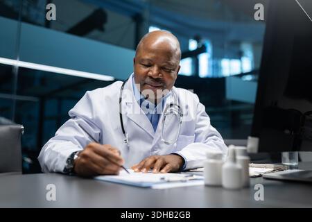 Docteur noir mature écrivant des notes médicales au bureau, documentant la paperasse Banque D'Images