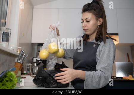 Une femme met des pommes jaunes d'un sac en plastique dans un récipient noir tout en se tenant debout dans sa cuisine. Elle porte un tablier et se concentre sur sa tâche, entourée d'outils de cuisine et de légumes. Banque D'Images