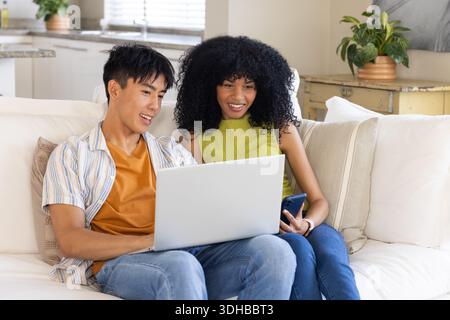 Couple diversifié partageant l'ordinateur portable argenté sur le canapé dans le salon, femme dans la citron vert tenant le smartphone Banque D'Images