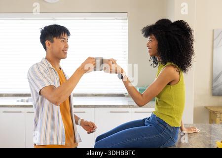 Homme asiatique et femme afro-américaine clinquant des tasses grises sur le compteur de quartz par stores, smartwatch Banque D'Images