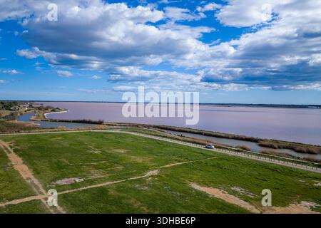 Lieu inconnu - 2 avril 2023 : L'image montre de vastes marais salants roses bordés de champs herbeux et une route avec une voiture garée sous un ciel partiellement nuageux. Banque D'Images