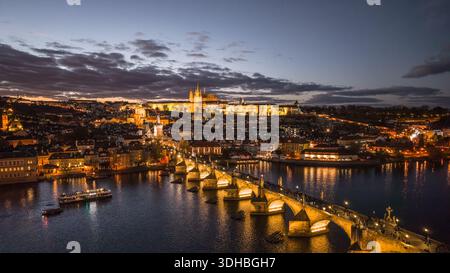 La scène capture le château de Prague et le pont Charles illuminés la nuit. La rivière Vltava reflète les lumières de la ville. Il montre une ligne d'horizon vibrante et une activité en soirée. Banque D'Images