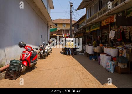 Kannur, Inde - 13 mars 2023 : une ruelle étroite avec des scooters garés le long d'un mur, des magasins ouverts vendant des marchandises dans de grands sacs, deux hommes debout près d'un pousse-pousse automobile, et une signalisation visible sous la lumière du jour claire. Banque D'Images