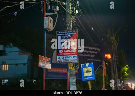Alappuzha, Inde - 8 mars 2023 : plusieurs panneaux de rue, y compris un panneau « No parking, Don't Drink and Drive » en anglais et en malayalam, un panneau « True Eyes » de la police du Kerala et un panneau d'arrêt de bus bleu sont attachés à un poteau avec des fils visibles la nuit. Banque D'Images