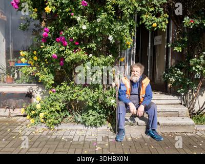 Un homme des années 70 avec une barbe blanche, assis sur des marches en béton devant un rosier en fleurs et une entrée de magasin Banque D'Images