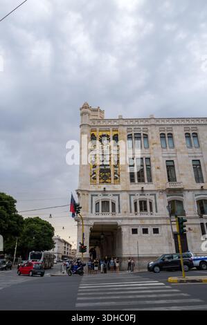 Cagliari, Italie - 20 septembre 2023 : des gens sont vus attendre à un passage devant un bâtiment historique en pierre arborant de grands armoiries et une façade décorative par temps nuageux. Banque D'Images