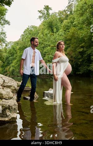 Couple enceinte tenant la main dans la nature marchant dans la rivière, au milieu de la forêt Banque D'Images