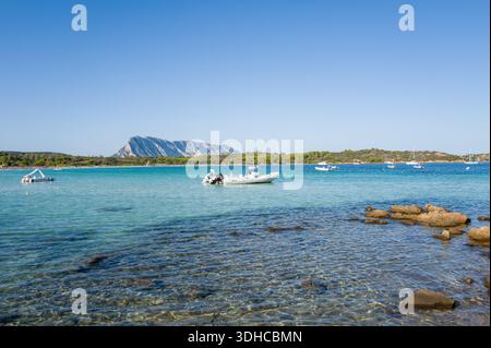 San Teodoro, Italie - 26 septembre 2023 : plusieurs bateaux à moteur et bateaux pneumatiques sont ancrés dans une eau claire peu profonde près d'un rivage rocheux à Spiaggia lu Impostu, avec une montagne lointaine et une végétation verte visible sous un ciel bleu clair. Banque D'Images