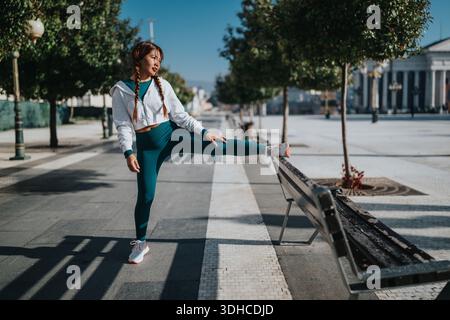 Femme athlétique en vêtements actifs sarcelle étire sa jambe sur un banc dans une place urbaine ensoleillée Banque D'Images