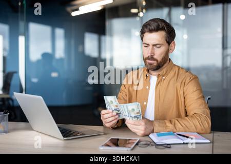 Homme avec la barbe assis à un bureau dans un bureau moderne, comptant avec précision une pile de billets de banque US dollar tout en gérant les finances et les tâches commerciales à l'aide d'un ordinateur portable personnel Banque D'Images