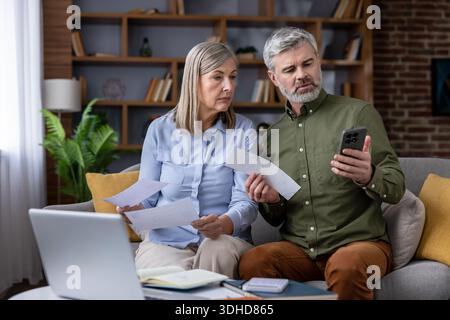 Couple d'âge mûr assis sur un canapé à la maison, analysant les finances personnelles, gérant le budget du ménage et examinant des documents avec un ordinateur portable et un smartphone pour les opérations bancaires en ligne Banque D'Images