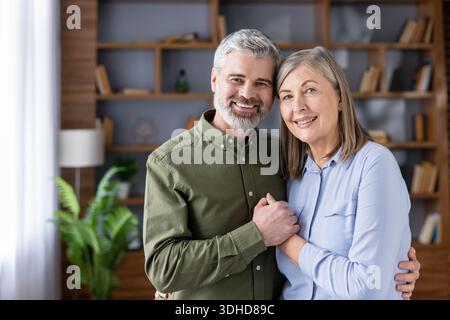 Mari et femme aînés aimant posant ensemble, embrassant et se tenant la main, exprimant le bonheur et la dévotion tout en regardant la caméra dans un salon moderne Banque D'Images