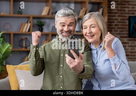 Sourire couple senior acclamant, célébrer les bonnes nouvelles et gagner un prix sur un téléphone portable tout en étant assis sur un canapé à la maison, sentir la joie et l'excitation Banque D'Images