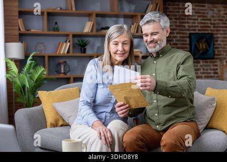 Couple plus âgé souriant assis sur un canapé dans leur salon, ouvrant une lettre d'une enveloppe brune, partageant un moment de convivialité et examinant des documents importants Banque D'Images