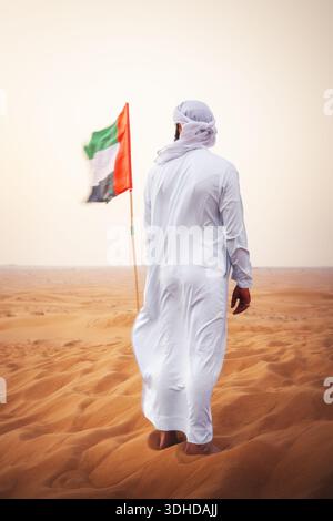 Vue d'un homme en vêtements blancs traditionnels se tient sur du sable doré ondulé, regardant vers le drapeau des Émirats arabes Unis sous un ciel pâle, Dubaï, Dubaï Banque D'Images