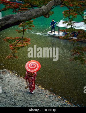 Kyoto, Japon - 18 janvier 2026 : vue d'une femme dans un kimono vibrant et un parasol rouge descendant vers la rivière, tandis qu'un batelier navigue dans le turquo Banque D'Images
