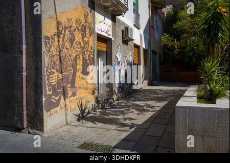 Orgosolo, Italie - 25 septembre 2023 : deux peintures murales sont peintes sur la façade d'un bâtiment le long d'une ruelle pavée, l'une montrant un groupe de personnes dans des tons sépia avec un texte italien manuscrit, et l'autre représentant plusieurs personnages en vêtements traditionnels, avec un petit arbre et une jardinière visibles au premier plan. Banque D'Images